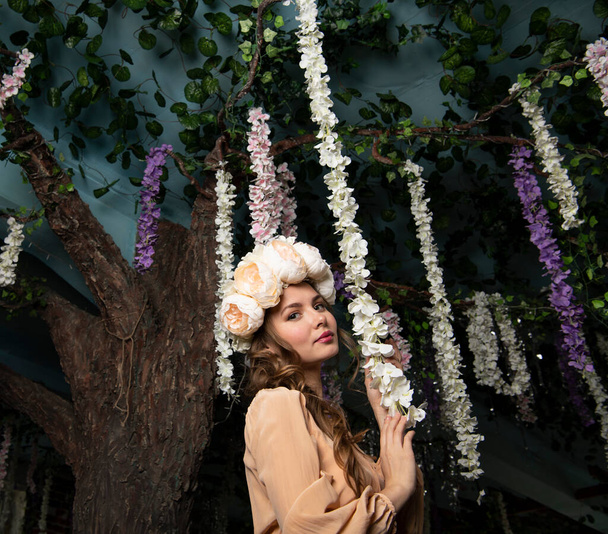 ανοιξιάτικο κορίτσι με ένα μπεζ φόρεμα και ένα λουλουδάτο στεφάνι στο κεφάλι της ποζάροντας σε φόντο λουλουδιών και δέντρων - Φωτογραφία, εικόνα