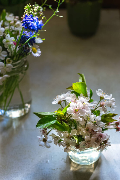 Small bouquets of cherry blossoms and spring field flowers in glass jars on old table. Closeup view, vertical orientation - Photo, Image