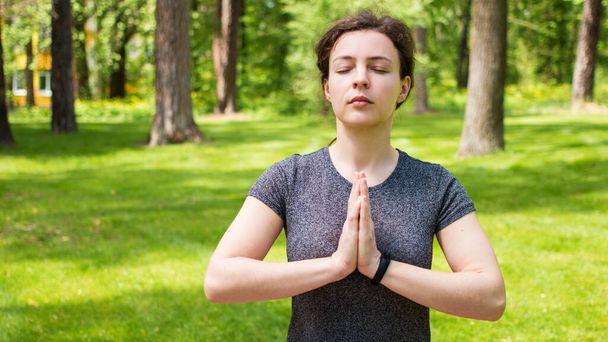 Молода спокійна жінка практикує йогу, розслабляючий медитуючий жест імен, вправляється в Падмасані. Жахлива мирна біла дівчина в сірій футболці, що практикує дихання йоги в зеленому парку на відкритому повітрі.
 - Фото, зображення