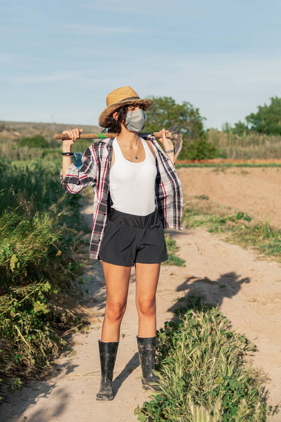 Молодая фермерша ходит в соломенной шляпе хирургическая маска и мотыга
 - Фото, изображение