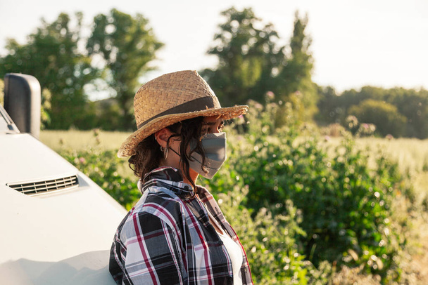 Jonge boerenvrouw met strohoed en chirurgisch masker leunend tegen het busje - Foto, afbeelding