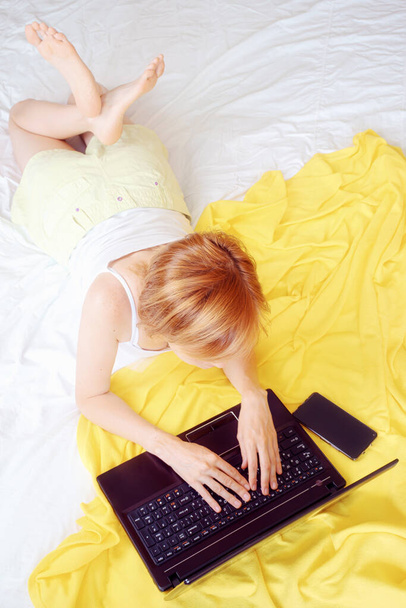 茶色の髪の白髪の少女は、ラップトップと入力してベッドの上に横たわっている。黄色のプレイ、スマートフォン。仕事の概念は、家庭で学ぶ。上からの眺め. - 写真・画像