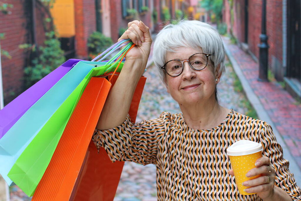 portrait of mature woman with grey hair in casual clothes with colorful paper shopping bags and paper cup of coffee on street - Zdjęcie, obraz