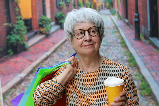 portrét dospělé ženy s šedými vlasy v ležérním oblečení s barevnými papírovými nákupními taškami a papírovým šálkem kávy na ulici - Fotografie, Obrázek