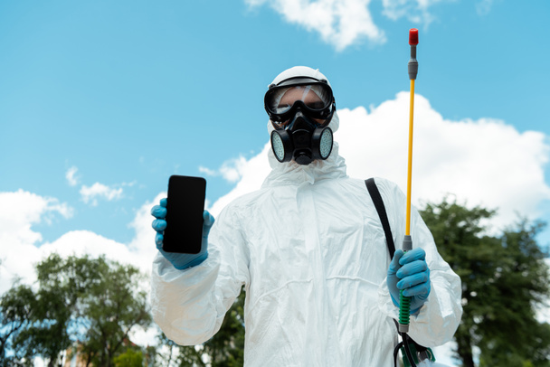 cleaning specialist in hazmat suit and respirator holding spray bag with disinfectant while showing smartphone with blank screen outdoors during coronavirus pandemic - Fotó, kép