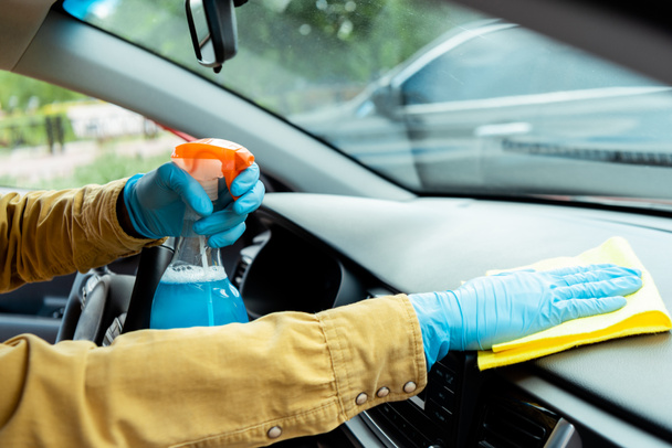 καλλιεργημένη άποψη του ανθρώπου με γάντια λατέξ χρησιμοποιώντας αντισηπτικό σπρέι και κουρέλι για τον καθαρισμό του εσωτερικού του αυτοκινήτου κατά τη διάρκεια της πανδημίας coronavirus - Φωτογραφία, εικόνα
