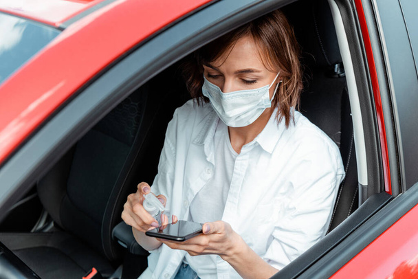 chica en máscara médica usando antiséptico para teléfono inteligente en el coche durante la pandemia covid-19
 - Foto, imagen