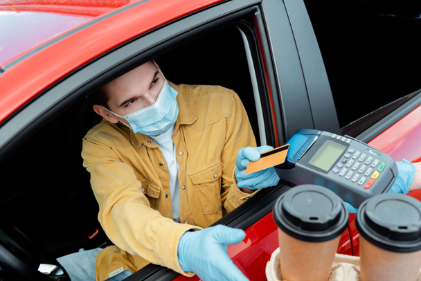 Mann in medizinischer Maske tätigt Transaktion mit Kreditkarte und Terminal, um während der Coronavirus-Pandemie Kaffee zum Mitnehmen aus dem Auto zu kaufen   - Foto, Bild