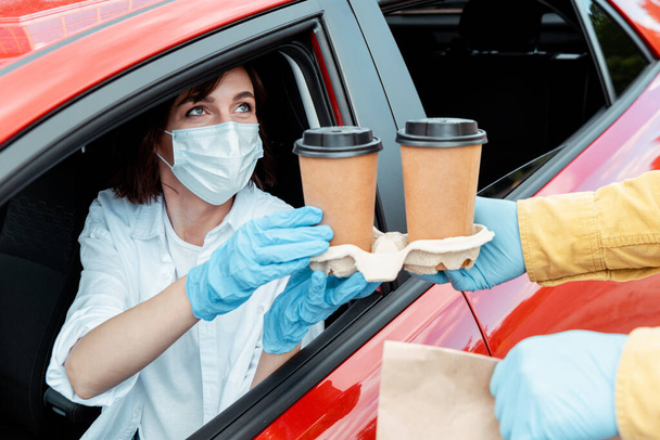 orvosi maszkos nő és kesztyű, aki kávét vásárol az autóból a koronavírus világjárvány idején   - Fotó, kép