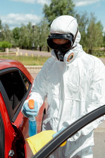 cleaner in hazmat suit cleaning car with antiseptic spray and rag during coronavirus pandemic - Photo, Image