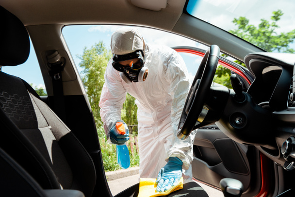 specialist in hazmat suit and respirator cleaning car interior with antiseptic spray and rag during coronavirus pandemic - 写真・画像