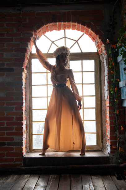 Bej elbiseli bir kız ve kafasında çiçekli bir çelenk pencerenin önünde poz veriyor. - Fotoğraf, Görsel