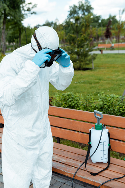 specialist in hazmat suit and respirator disinfecting bench in park during coronavirus pandemic - Foto, afbeelding