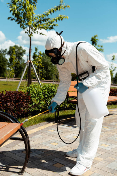 specialist in hazmat suit and respirator disinfecting bench in park during coronavirus pandemic - 写真・画像