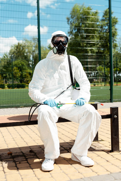 specialist in hazmat suit and respirator sitting on bench with spray bag during covid-19 pandemic - Фото, изображение