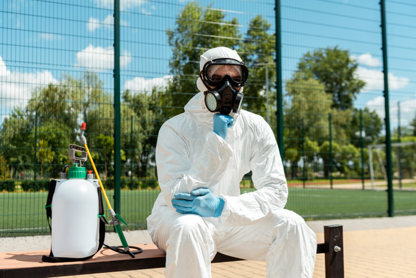 especialista en limpieza en traje de materiales peligrosos y respirador pensando y sentado en el banco con bolsa de spray durante la pandemia de covid-19
 - Foto, Imagen