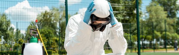 preoccupato specialista in hazmat suit e respiratore seduto su panca con borsa spray durante la pandemia covid-19, orientamento panoramico
 - Foto, immagini