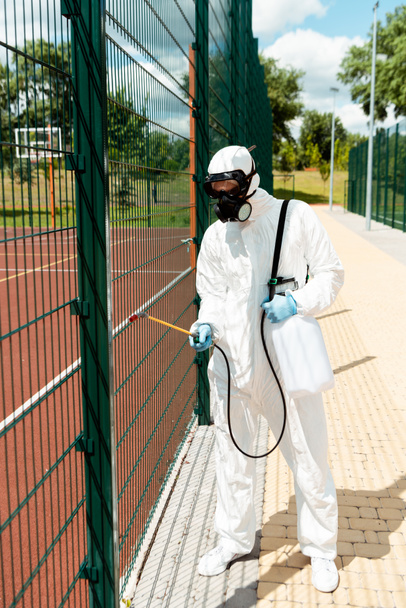 professional specialist in hazmat suit and respirator disinfecting fence of basketball court in park during coronavirus pandemic - Photo, image