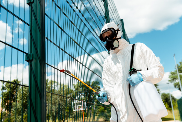 male specialist in hazmat suit and respirator disinfecting fence of basketball court in park during covid-19 pandemic - Photo, Image