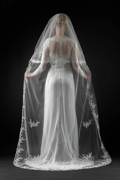 Bride in a beaituful flowing white wedding dress and long lace bridal veil, back view. Black background - Photo, image