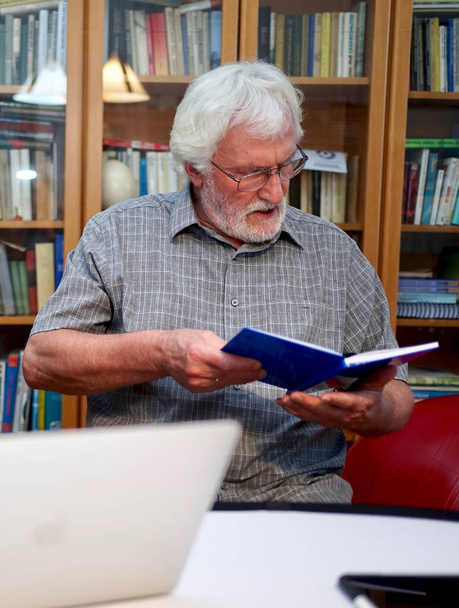 Retrato de um homem idoso barbudo branco com camisa cinza lendo um caderno azul, livro enquanto sentado na frente de uma prateleira de livro enorme no fundo
.   - Foto, Imagem