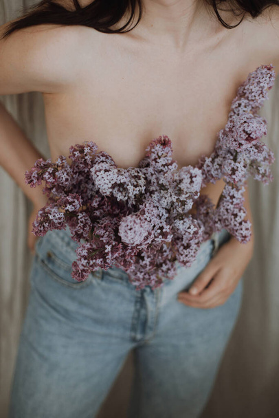 Blooming lilac flowers covering naked woman upper body on rustic background. Woman posing with lilac branches in denim jeans. Creative moody image. Sensuality and tenderness concept - Photo, Image