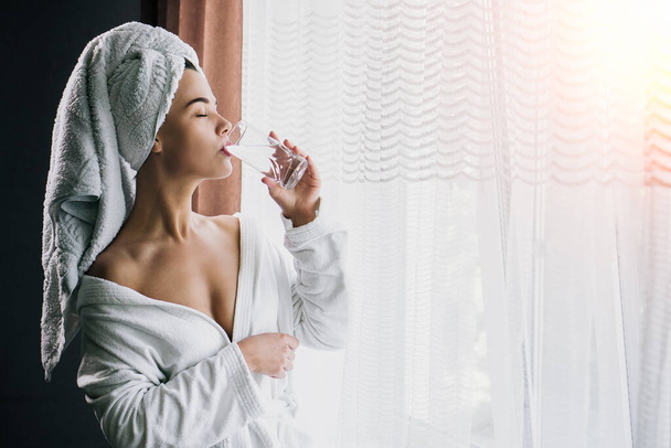 Nuori kaunis nainen valkoisessa pyyhkeessä ja kylpytakki juomaveden lähellä ikkunan kotona
 - Valokuva, kuva
