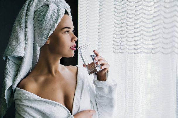 Jovem mulher bonita em toalha branca e manto água potável perto da janela em casa
 - Foto, Imagem