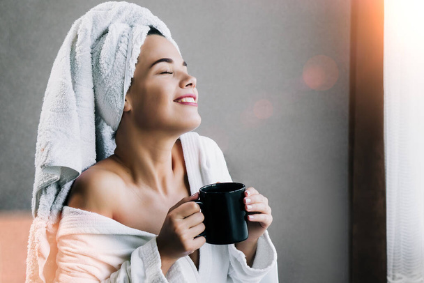Young beautiful woman in white towel and robe drinking coffee and enjoying near the window at home - Photo, Image
