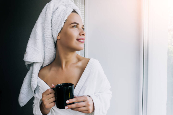 Retrato de jovem mulher bonita em toalha branca e roupão beber café e desfrutar perto da janela em casa
 - Foto, Imagem