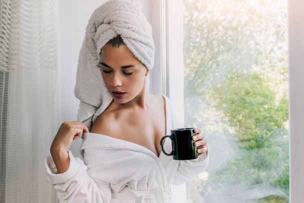 Fiatal gyönyörű nő fehér törölközőben és köntösben kávézik az ablak mellett otthon. - Fotó, kép