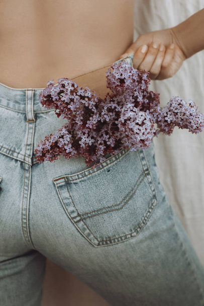 Blooming lilac flowers in back pocket. Woman posing with lilac flowers in denim jeans pocket on rustic background.  Creative image. Sensual atmospheric mood. Hello spring - Foto, immagini