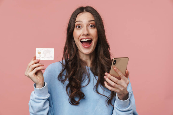 Image of attractive young woman with long brown hair rejoicing while holding cellphone and credit card isolated over pink background - Foto, Bild