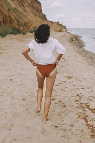 Chica boho en camisa blanca caminando por la playa soleada, vista trasera. Mujer con estilo sin preocupaciones en traje de baño y camisa relajante en la orilla del mar. Vacaciones de verano. Estilo de vida imagen auténtica
 - Foto, imagen