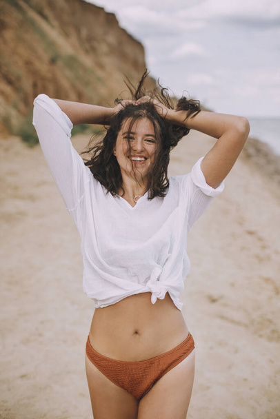 Joyful boho girl in white shirt having fun on sunny beach. Carefree stylish woman in swimsuit and shirt smiling and relaxing on seashore. Summer vacation. Lifestyle image - Foto, imagen