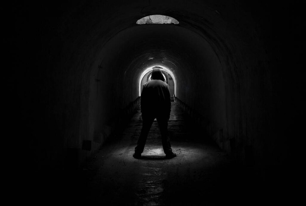 La figura de un hombre en una capucha en un oscuro túnel subterráneo sombrío, de pie frente a la luz e iluminado a través de un agujero en el techo
. - Foto, Imagen