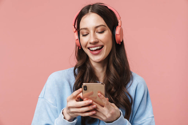 Image of delighted young woman laughing while using headphones and cellphone isolated over pink background - Foto, imagen