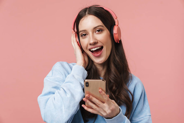 Image of delighted young woman laughing while using headphones and cellphone isolated over pink background - Fotó, kép