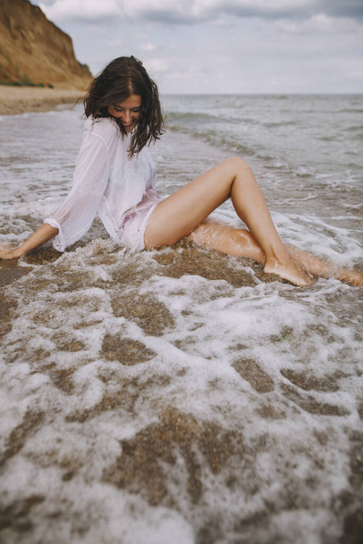 Szczęśliwa kobieta w białej koszuli siedząca na plaży w rozbryzganych falach. Stylowa opalona dziewczyna relaksująca się na wybrzeżu i ciesząca się falami. Wakacje. Uważność i beztroska chwila - Zdjęcie, obraz