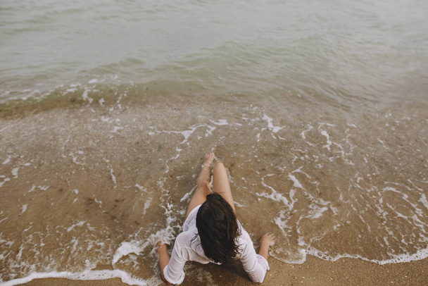 Young woman in wet white shirt lying on beach in splashing waves. Top view. Stylish tanned girl relaxing on seashore and enjoying waves. Summer vacation. Mindfulness and carefree - Photo, Image