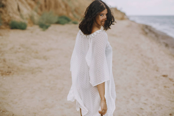 Beztroska dziewczyna boho w białej letniej sukience spacerująca po plaży. Szczęśliwa młoda kobieta relaksująca się na wybrzeżu. Wakacje. Uważność i relaks. Styl życia - Zdjęcie, obraz