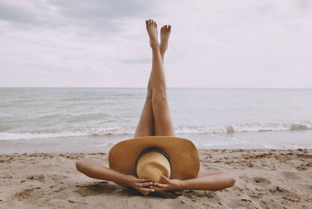 Wakacje i podróże. Dziewczyna w kapeluszu leży na plaży z nogami w górze. Modna młoda kobieta trzymająca słomkowy kapelusz, relaksująca się na piaszczystej plaży w pobliżu morza. Beztroski obraz kreatywny - Zdjęcie, obraz