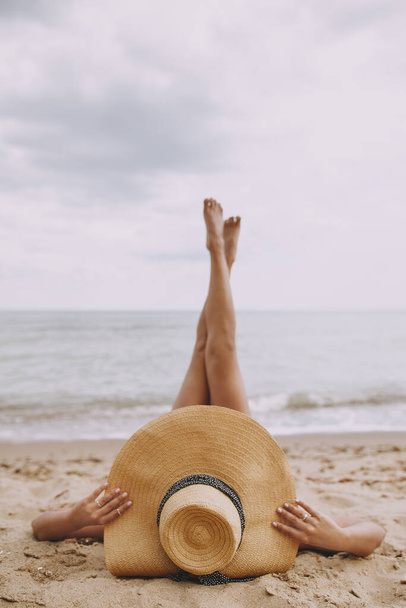 Літні канікули та подорожі. Дівчина в капелюсі лежить на пляжі з ногами вгору. Модна молода жінка тримає солом'яний капелюх, розслабляючись на піщаному пляжі біля моря. Безтурботний креативний образ
 - Фото, зображення