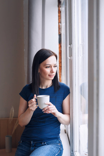 Beautiful woman at home drinking a cup of coffee and looking at window - Фото, изображение