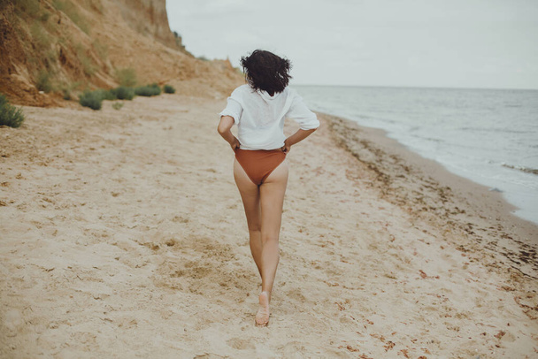 Boho dziewczyna w białej koszuli działa na słonecznej plaży, widok z tyłu. Beztroska stylowa kobieta w stroju kąpielowym i koszuli relaksująca się na brzegu morza. Wakacje. Autentyczny wizerunek stylu życia - Zdjęcie, obraz
