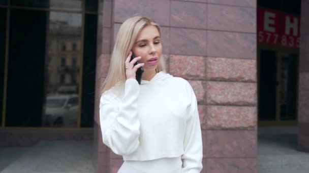 portrait of a young attractive blonde woman in a white suit standing smiling talking on the phone. is having a conversation. Hipster student girl in love. urban street background. - Footage, Video