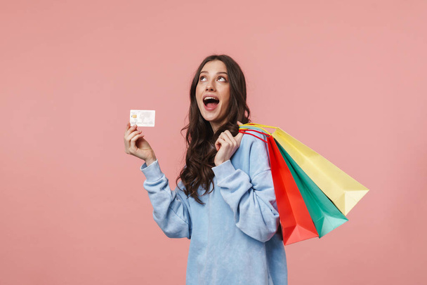 Image of attractive young woman with long brown hair holding shopping bags and credit card isolated over pink background - Photo, image