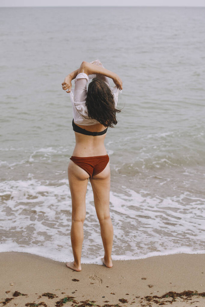 Onnellinen nuori nainen ottaa pois märkä valkoinen paita rannalla, takaisin näkymä. Tyylikäs tyttö uimapuku rentouttava meren rannalla. Kesäloma. Huoleton hetki
 - Valokuva, kuva