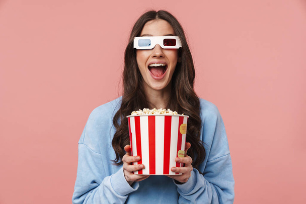 Portrait of a pretty cheerful girl with long curly brunette hair wearing sweatshirt standing isolated over pink background, wearing 3d glasses, holding popcorn bucket - Foto, Imagen