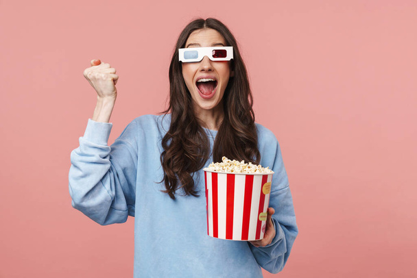 Portrait of a pretty cheerful girl with long curly brunette hair wearing sweatshirt standing isolated over pink background, wearing 3d glasses, holding popcorn bucket, celebrating - Fotó, kép